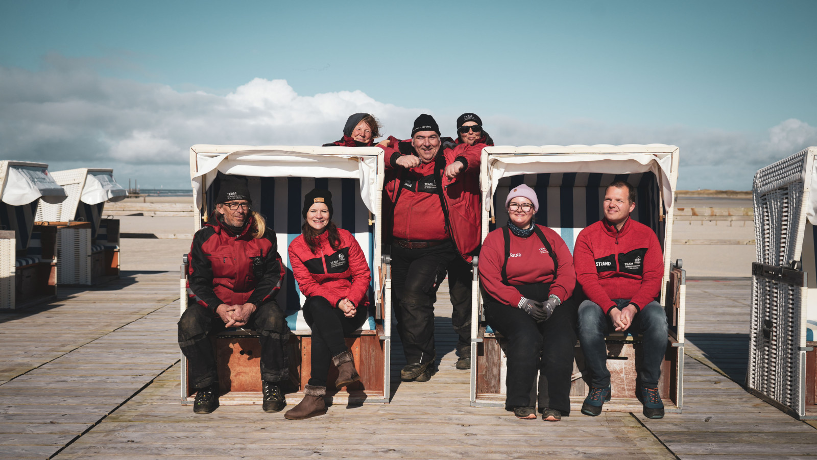 Mehrere Personen sitzen und stehen in und neben Strandk&ouml;rbern auf einem Podest in St. Peter-Ording. Im Hintergrund wei&szlig;er Sandstrand