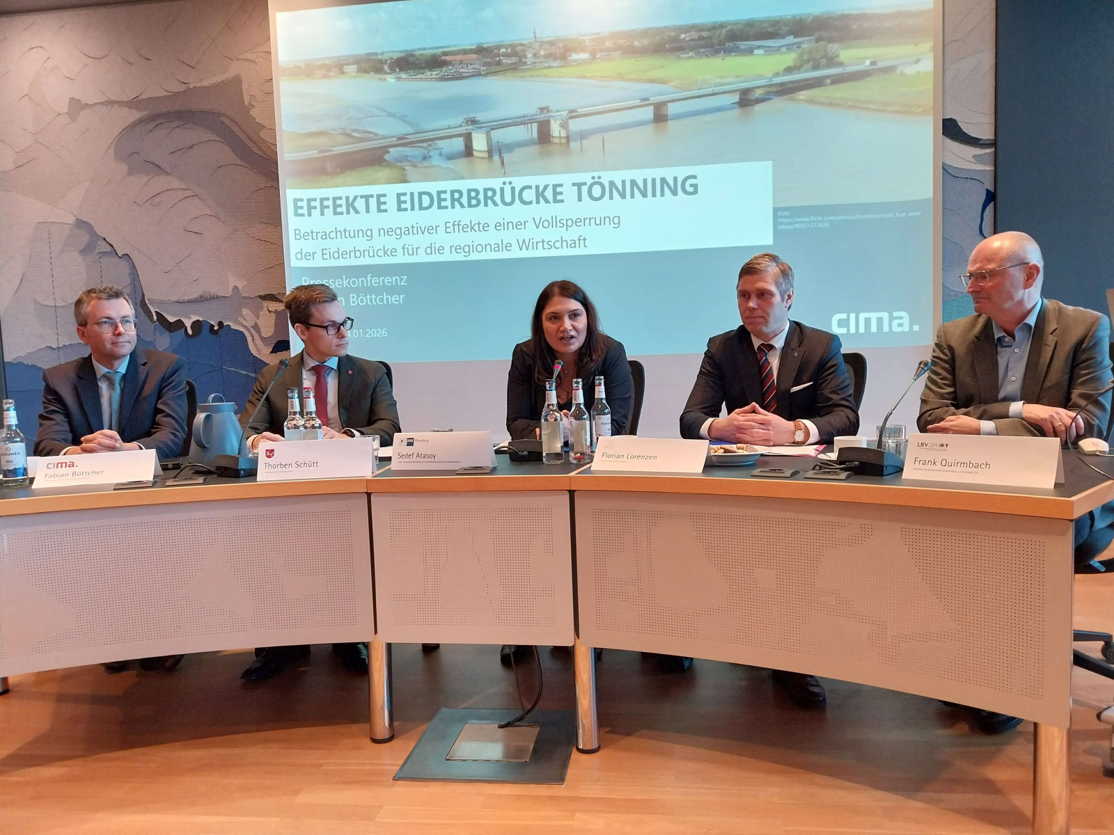 F&uuml;nf Personen sitzen an einem gebogenen Konferenztisch vor einer Pr&auml;sentation mit dem Titel 'Effekte Eiderbr&uuml;cke T&ouml;nning'.