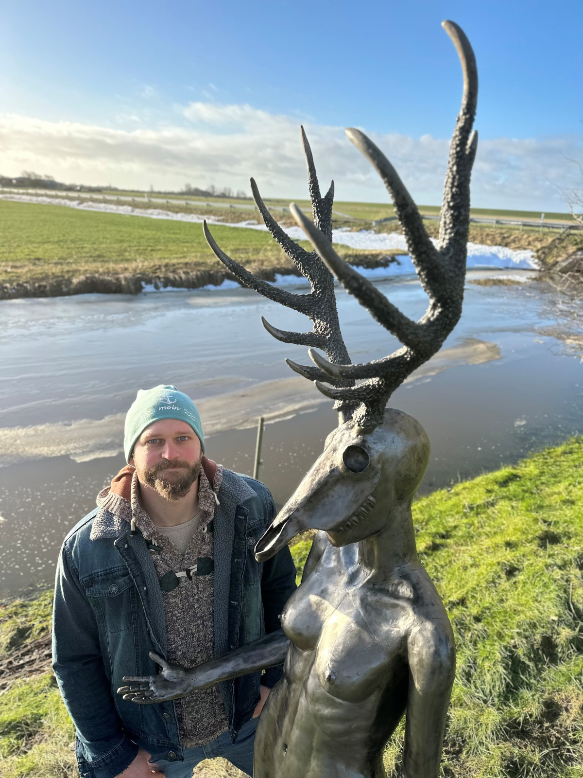 Bronzefigur mit Hirschgeweih und menschlichem K&ouml;rper steht an einem Flussufer neben einer Person in Jeansjacke