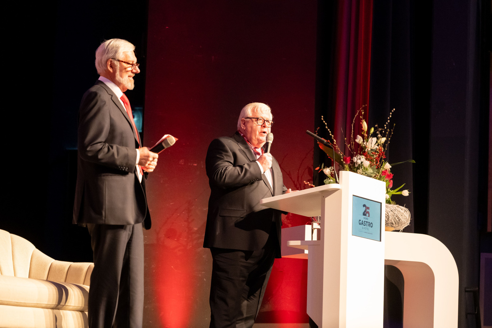 Zwei M&auml;nner in Anz&uuml;gen stehen auf einer B&uuml;hne, einer spricht am Rednerpult mit Blumenarrangement und Logo mit Schriftzug 'GASTRO'